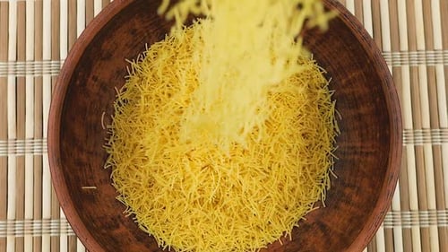 Thin small pasta is poured into a brown ceramic bowl against a bamboo mat. Top view. Slow motion.