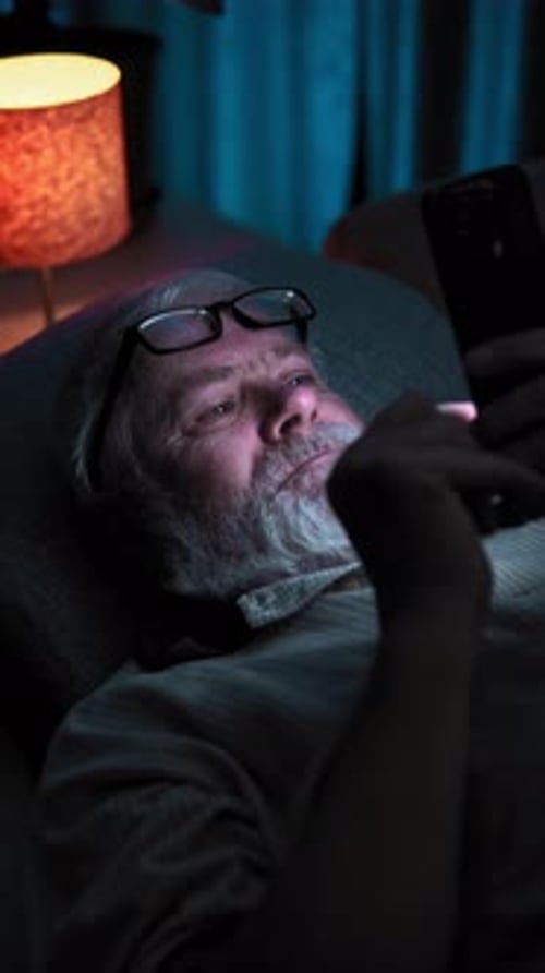 Elderly Man with Glasses Lies in Bed and Surfing News on His Smartphone