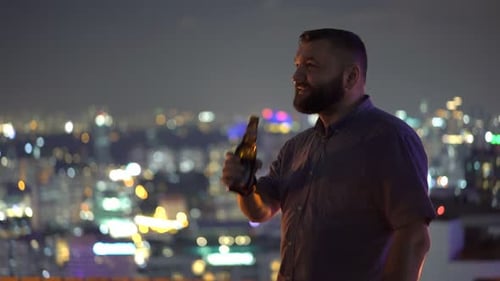 Handsome man enjoys a cold beer and city lights view from rooftop skybar at night