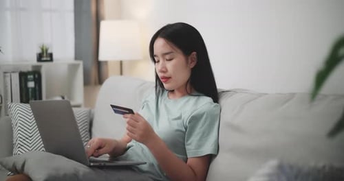 Young woman uses laptop and credit card to shop online on sofa at home.