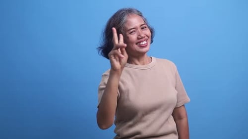 Middle age Asian woman standing and smiling showing two fingers in winning gesture, victory sign, ov