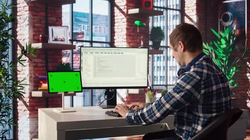 Man in Home Office Looking at Green Screen Tablet and Computer Typing Code