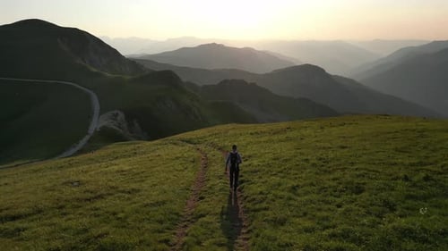 A man with a backpack walks along a green mountain path at sunset. Aerial view.