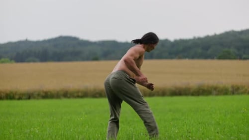 Shirtless Man is Practicing Martial Arts in a Tranquil Grassy Field Against a Rural Landscape