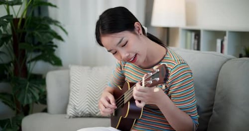Beautiful young woman plays ukulele and sings in living room at home during holiday.