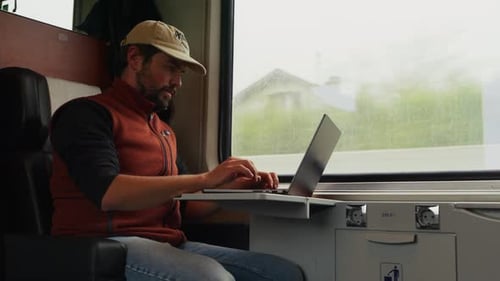 Ein Mann sitzt am Zugfenster und konzentriert sich auf seinen Laptop, während draußen die Szenerie vorbeirauscht -