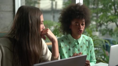 Two young diverse female colleagues working at coffee shop in front of laptop computer