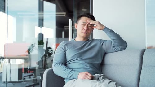 Young exhausted, overworked office worker sitting on a couch with a severe headache massaging