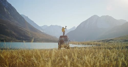 Photographer Standing on SUV Roof with Camera By Mountain Lake