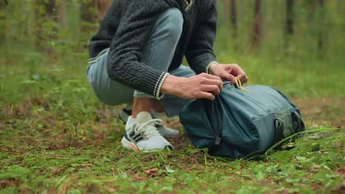 Individual Squatting in Forest Unzipping Camping Bag with Partial Gear Visible