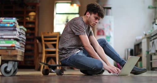 Young Man Uses Laptop Sitting on Skateboard Indoors