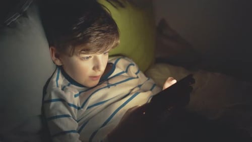 Un garçon souriant heureux est allongé dans son lit sur des coussins, des émotions, joue sur un smartphone, un jeu sombre