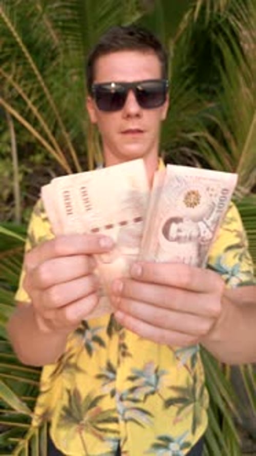 Man Counting Thai Baht on a Beach with Palm Leaves in the Background