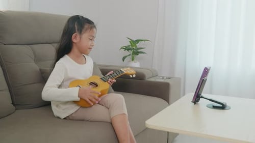 A Young Girl Sits on a Couch Playing a Ukulele with a Smartphone on a Stand Streaming Her Online