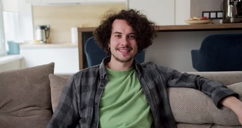 Porträt eines glücklichen brünetten Mannes mit lockigem Haar, Schnurrbart und kariertem Hemd, der auf dem Sofa sitzt
