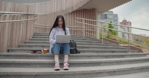 Joven estudiante asiática que usa una computadora portátil mientras está sentada en una escalera al aire libre