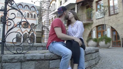 Pareja sentada en un muro de piedra entre los edificios de la plaza del pueblo y besándose, SLOMO