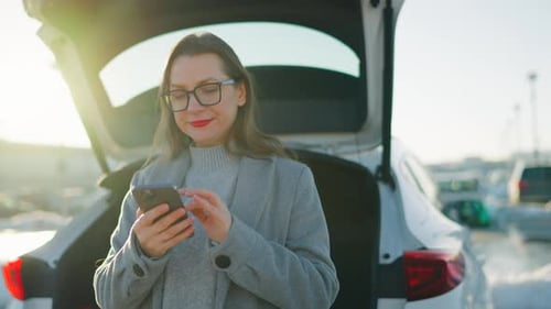 Woman Uses Phone By Car Trunk in City