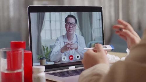 Close Up Of Sick Asian Businesswoman Holding A Thermometer To The Video Call With Doctor On A Laptop