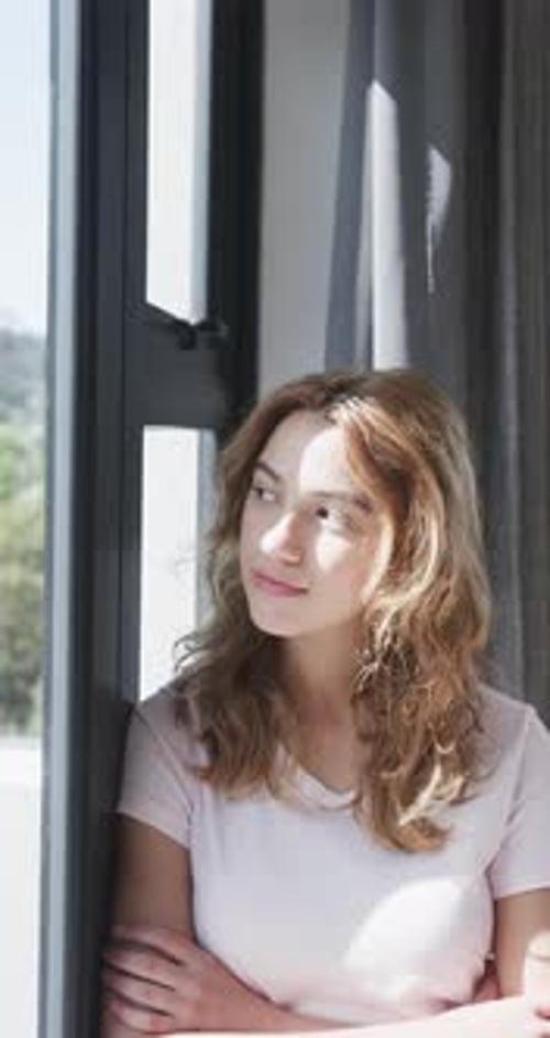 Happy caucasian woman looking by window and smiling in sunny house