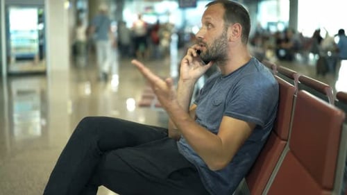 Frustrated man on cellphone awaiting delayed flight in busy airport terminal