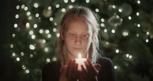 The Child Holds a Burning Candle in His Hands Against the Backdrop of Blurry Christmas Tree Lights