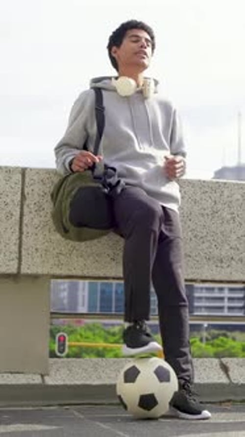 Vertical video: Balancing man in hoodie tapping ball on ledge after friend arrival, holding duffel