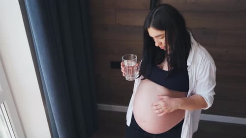 Eine schwangere Frau, eine werdende Mutter, steht am Fenster und trinkt Wasser, Gesundheit und Schwangerschaft