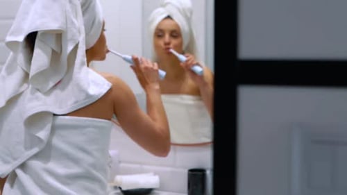 Linda chica caucásica con una toalla blanca en la cabeza y el cuerpo se cepilla los dientes con un eléctrico