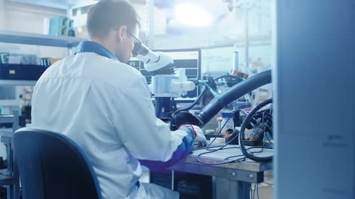 Electronics Factory Worker in White Work Coat Inspects a Printed Circuit Board Through a Digital Mi