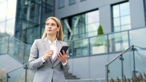 Confident businessman is using a phone while walking on the street near an office building. Serious