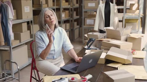 Mature woman fashion dropshipping business owner talks on phone in warehouse