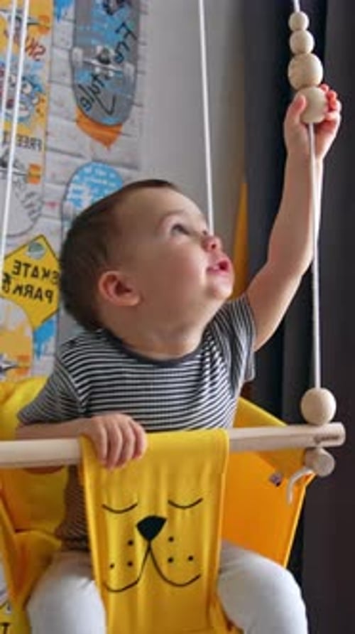 Little boy swings in a yellow swing indoors. Kid plays with wooden beads on the rope. Vertical video