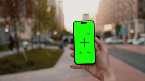 Closeup of a Hand Holding a Smartphone with a Mockup Green Screen with a City Street on a Sunny Day