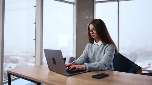 Long-haired brunette sits at desk typing on her laptop. Businesswoman working in the spacious office