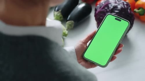 Modern Smartphone With Green Screen For Chroma Key Montage In Hand Of Mature Woman in Kitchen