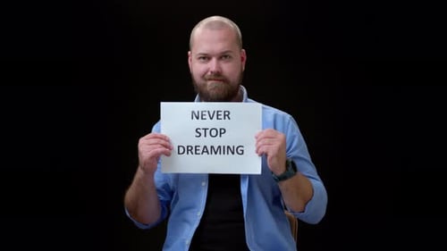 Bald Bearded Man in Blue Shirt Sit Against Black Background Holding Up White Sheet of Paper That