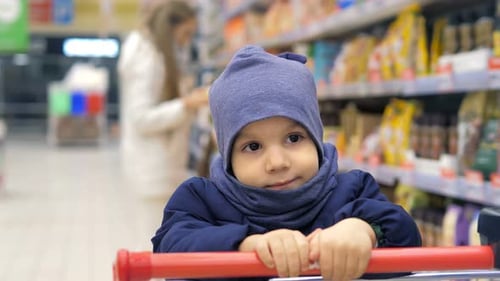 Young Mother is Choosing Food in Supermarket on the Background of Very Sad 3 Years Old Boy Sitting