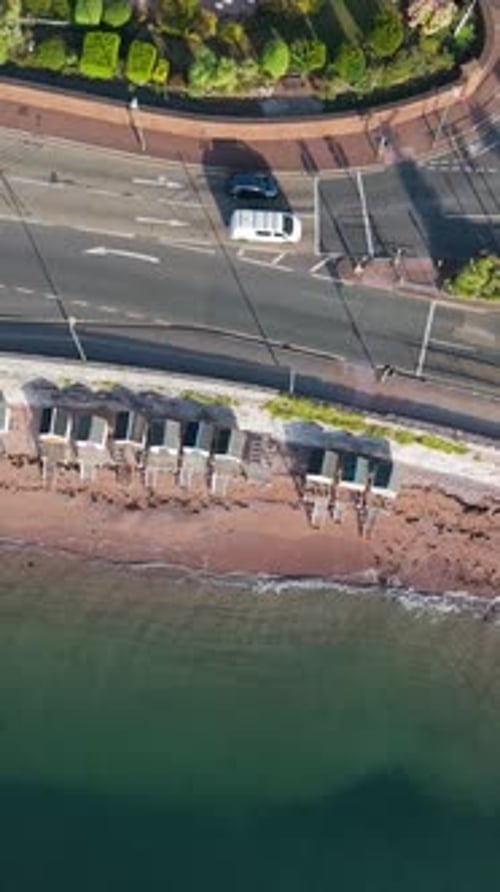 Vertical top-down drone view over Torquay beach huts, blue sea, waves, road and coastline from above