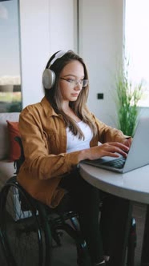 Woman in Wheelchair Using Laptop at a Table in a Modern Room with Plants While Wearing Headphones