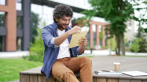 Smiling male student reading letter with great news sitting on a bench in campus near university