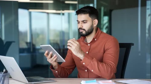 Thoughtful businessman is using digital tablet sitting at workplace in business office. Handsome