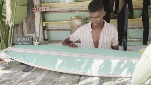 African american man preparing surfboard on the counter of surf rental beach shack, slow motion