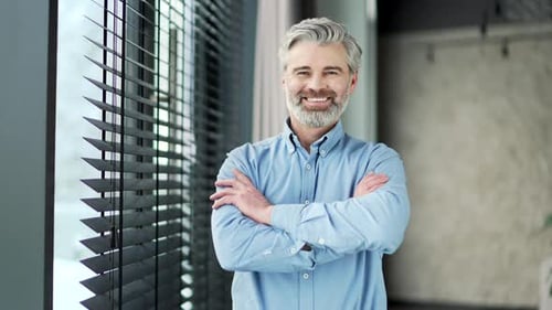 Portrait of mature gray haired bearded businessman standing with crossed arms in business office.