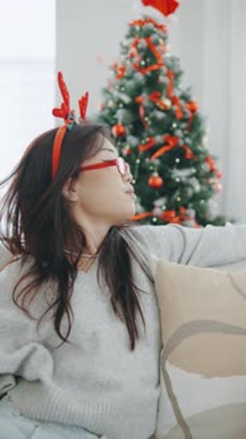 Young Woman with Reindeer Headband Sitting on a Sofa Near a Decorated Christmas Tree During Holiday