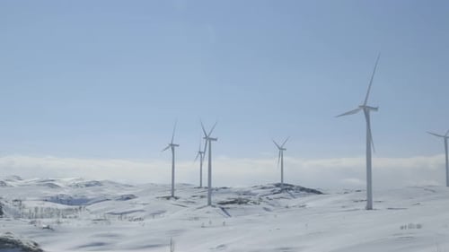 Die Kamera schwenkt nach links über verschneite Berge, in denen sich Windräder unter hellem Himmel stetig drehen.