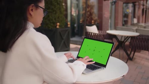 Young Woman Typing on Laptop with Green Screen at Outdoor Cafe
