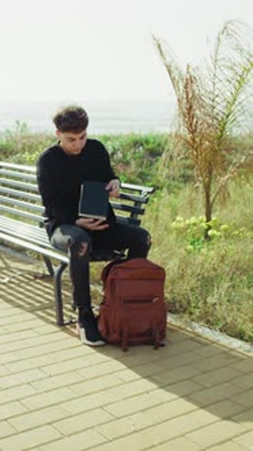 Young Man On A Bench Near The Sea Picking A Notebook From The Backpack