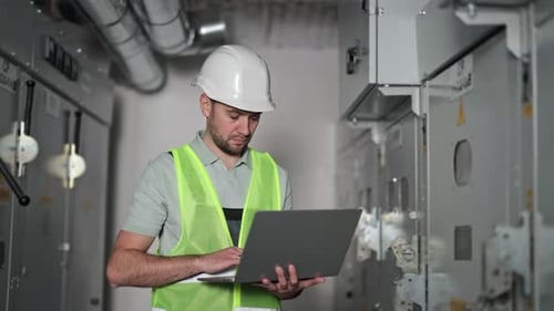 Electrical Engineer Working on Laptop at Electrical Control Panel in Substation