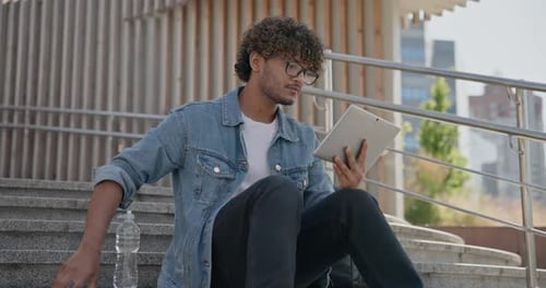 Jeune bel homme indien hindou étudiant indépendant assis à l'extérieur sur des marches à l'aide d'une tablette PC